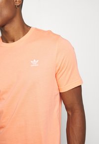 Muž v korálovém tričku Adidas s bílým logem na hrudi, ukazující levou stranu horní části těla a paže s malými kruhovými náušnicemi.