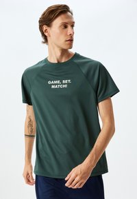 Mörkgrön sportt-shirt i andningsbart material. Har raglanärmar och vit text "GAME, SET, MATCH!" på framsidan.