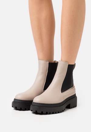 Beige læderankelstøvler med sorte elastiske sidepaneler, chunky sort sål og glat tekstur. Har minimal syning og rundet tå.