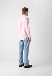 Gaiši rozā krekls ar garām piedurknēm un izliekta apakša, apvienots ar gaiši ziliem džinsiem un melnām kurpēm, skatīts no aizmugures.