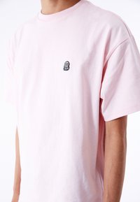Camiseta de algodón rosa claro con un corte relajado. Presenta un pequeño logotipo negro bordado en el pecho. Mangas cortas y cuello redondo.