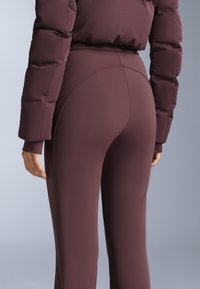 Bordeauxrode sportleggings met een hoge taille en gebogen naaddetails. Gemaakt van een glad, rekbaar materiaal; gecombineerd met een gewatteerd jack.