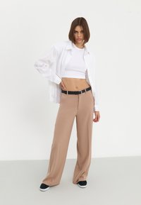 Pantalones de pierna ancha beige con una textura suave, combinados con un top corto blanco y una camisa blanca de gran tamaño. Un cinturón negro acentúa el conjunto.