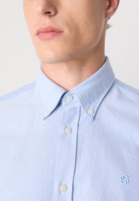 Šviesiai mėlynas mygtukų marškiniai pagaminti iš medvilnės ir sintetinių audinių mišinio. Turintys aštrų apykaklę, baltas mygtukų akcentus ir logotipą ant krūtinės. Lygus paviršius.