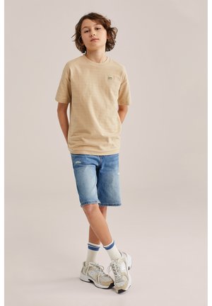 Poiss seisab käed selja taga, seljas beež t-särk, teksapüksid, valged sokid siniste triipudega ja valged tossud neutraalsel taustal.