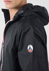Uomo che indossa una giacca nera con cappuccio, con una cerniera rossa e bianca; sul maniche è presente un'etichetta con il logo JOTT che mostra una silhouette su scooter.