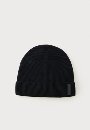 Bonnet en tricot noir avec bord rabattu et petite étiquette rectangulaire sur le côté, présenté sur un fond clair uni.