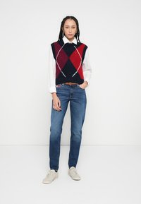 Modrý a červený argyle pletený sveter bez rukávov cez bielu košeľu s golierom, kombinovaný s modrými džínsy a svetlými teniskami. Jednoduchý, neformálny outfit.