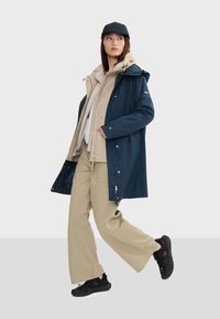Manteau imperméable marine superposé sur une veste beige, un pantalon beige et des chaussures noires. Il comprend une capuche, des zippers et des boutons-pression.