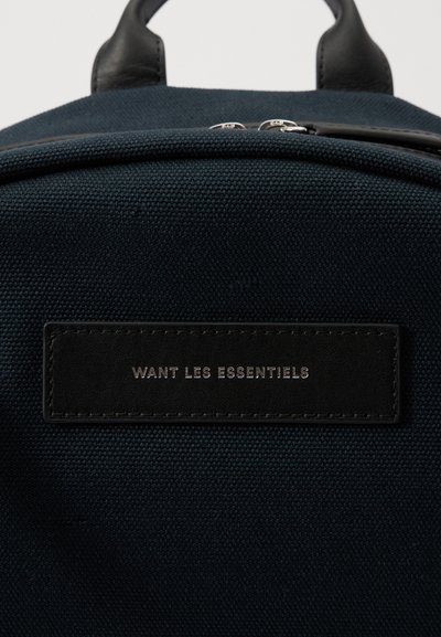 Sötét tengerészkék vászon hátizsák fekete bőrcímmel, amelyen az "WANT LES ESSENTIELS" felirat található, dombornyomott betűkkel. Simább tapintás, cipzáras zárás, letisztult dizájn.