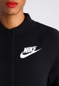 Černá mikina s vysokým límcem a raglánovými rukávy. Na levé straně je velké bíl�é logo Nike. Bavlněný materiál s hladkou texturou.