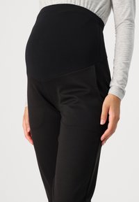 Zwarte zwangerschapsbroek van een zachte, rekbare stof met een hoge, elastische tailleband. Kenmerkt zich door een gladde textuur en een getailleerd ontwerp.