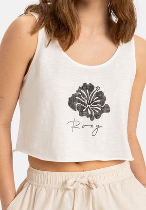Kobieta nosząca biały krótki top z czarnym kwiatem hibiskusa i napisem „Roxy”, w połączeniu z beżowymi spodniami ze sznurkiem.