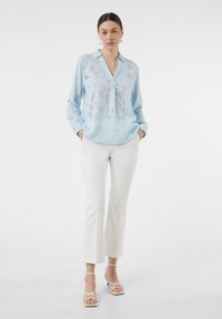 Helles Blaues Bluse mit einem subtilen transparenten Überzug, mit Kragen und langen Ärmeln. Kombiniert mit weißen Slim-Fit-Hosen und sandalen mit Riemchen.