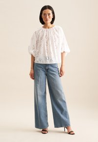 Top bianco ricamato a occhio di pernice con scollatura arricciata, abbinato a jeans blu in denim a gamba larga e sandali neri con tacco. Tessuto morbido e fluido.