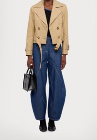 Beige cropped trenchcoat met dubbelzijdige knopen, gecombineerd met donkerblauwe oversized denim jeans met contrasterende stiksels, en een kleine zwarte handtas.