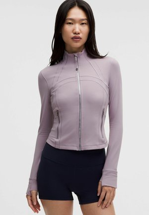 Kvinde med langt mørkt hår iført en tætsiddende lavendel-lilla lynlåstrøje og sorte shorts, stående mod en ensartet lys baggrund.