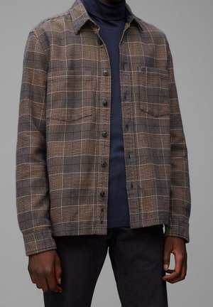 Homme portant une chemise à carreaux marron boutonnée sur un col roulé bleu marine foncé avec un pantalon noir, debout devant un fond gris neutre.