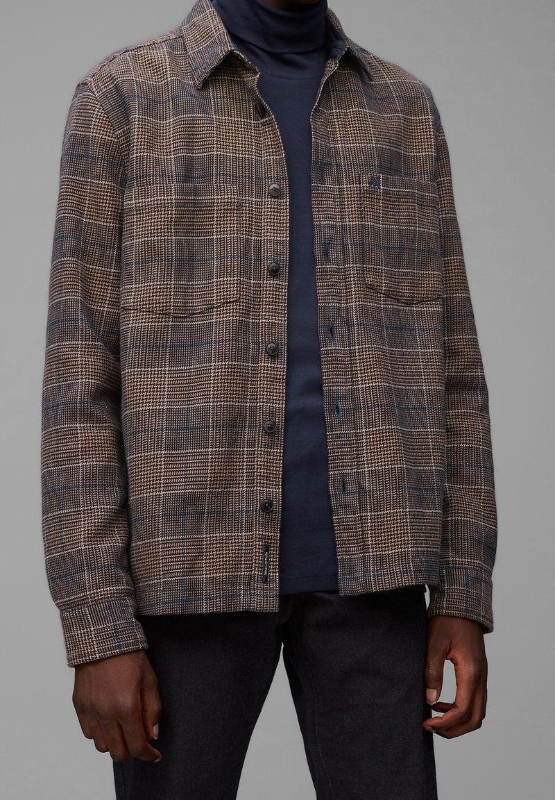 Homme portant une chemise à carreaux marron boutonnée sur un col roulé bleu marine foncé avec un pantalon noir, debout devant un fond gris neutre.