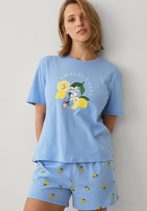 Kvinde iført lyseblåt afslapningstøj med citron- og blomsterdesigns samt teksten "Amalfi Coast", stående foran en ensfarvet hvid baggrund.