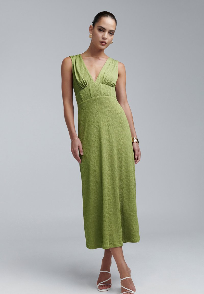 Groene, getextureerde, mouwloze jurk met een diepe V-hals en geplooide taille. Valt tot halverwege de kuit, gecombineerd met witte sandalen met bandjes.