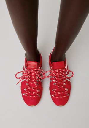 Persona de piel oscura que lleva zapatillas deportivas rojas brillantes con rayas plateadas y cordones con estampado, de pie sobre un suelo gris claro.
