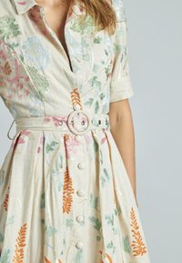 Abito in lino beige con ricami floreali, dotato di maniche corte, una vita cintrata con una fibbia tonda e dettagli con bottoni sul davanti.