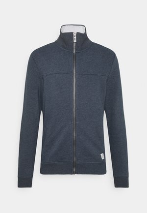 Tume sinine lukuga jakk kõrge kaelusega, soonikute ja servadega. Valmistatud pehmest materjalist, millel on kerge tekstuur ja väike logo allosas.