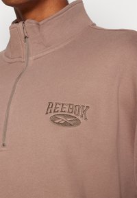 Světlý fialkový pulovr Reebok s čtvrtým zipem, texturovaným povrchem, vyšitým logem a vysokým límcem pro teplo.