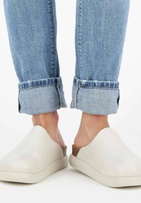 Personne portant un jean bleu clair retroussé et des chaussures beiges à enfiler sans dossier, se tenant sur une surface blanche, les talons exposés.