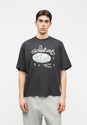 Νεαρός άνδρας με κοντά σκούρα μαλλιά, που φοράει μια μαύρη, υπερμεγέθη μπλούζα με χάρτη του κόσμου και το λογότυπο "adidas", συνδυασμένη με ανοιχτό γκρι παντελόνι.