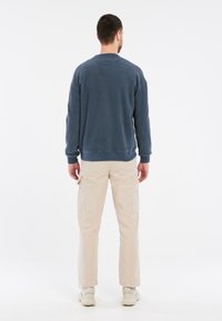 Felpa blu navy con collo a giro e polsini a coste, abbinata a pantaloni cargo beige con tasche laterali. Le scarpe sono di colore chiaro.