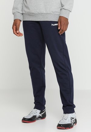 Man draagt marineblauwe hummel joggingbroek, grijze trui en wit-zwarte-rode sportschoenen, staand op een effen achtergrond.