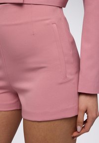 Pantaloni corti su misura di un delicato colore rosa, realizzati in un tessuto liscio, con tasche laterali e un orlo pulito, mostrati di lato con una mano appoggiata sul tessuto.