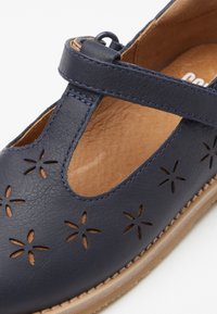 Granatowe skórzane buty z wyciętym kwiatowym wzorem, z paskiem T i beżową podeszwą. Gładka tekstura z kontrastującymi ciemnymi szwami.