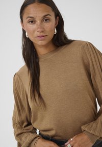 Pull en maille côtelée marron avec des manches longues bouffantes et un col rond. Le modèle porte une ceinture noire et des boucles d'oreilles en argent. Texture lisse.