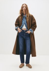Hnedý oversized kabát cez džínovú bundu, biely top a modré džínsy. Obsahuje svetlohnedé mokasíny, ktoré pridávajú textúru outfitu.