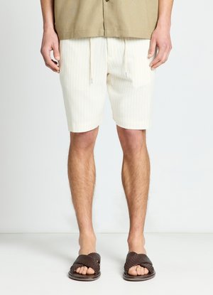 Mann trägt beige Shorts mit Kordelzug, braunes Oberteil und braune gewebte Sandalen und steht vor einem einfarbigen hellen Hintergrund.