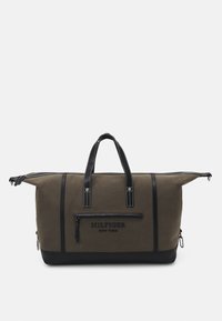 Sac de voyage en toile vert olive avec détails en cuir noir, double poignées, fermeture éclair, poche avant et coutures blanches.
