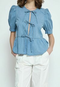Top in denim azzurro chiaro con maniche a puff corte, dettagli a fiocco sul petto e orlo a peplo. Indossato con pantaloni cargo bianchi larghi.
