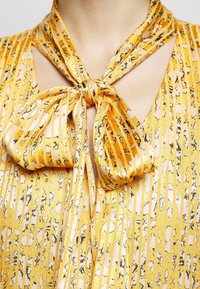 Blouse jaune à motifs avec des rayures verticales, dotée d'un col noué. Le tissu a une texture lisse avec un léger éclat.