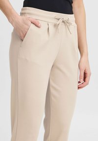 Beige joggingspakken gemaakt van zacht materiaal, met een elastische tailleband, verstelbaar trekkoord en zijzakken. Gladde textuur met strakke lijnen.