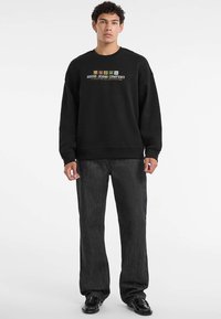 Męski model w czarnej bluzie z kapturem z logo "Guess Jeans Company", w ciemnoszarych, luźnych dżinsach i czarnych, wypolerowanych butach wizytowych, stojący na białym tle.