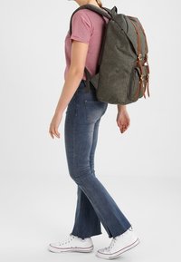 Mujer vestida con una camiseta de rayas rojas, jeans de campana azules, zapatillas blancas y llevando una gran mochila gris con correas de cuero marrón.