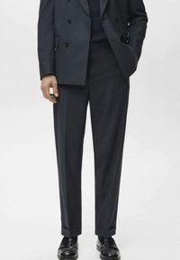 Pantalon tailleur marine en tissu lisse, présentant une coupe droite et un pli avant discret. Porté avec des chaussures noires.