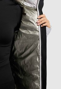 Černá vnější textilie bundy, otevřená tak, aby odhalila stříbrné podšívku s krčenou texturou. Obsahuje kontrastní bílou vnitřní část a viditelné švy.