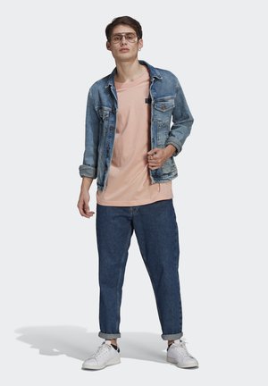 Giacca di denim sopra una t-shirt rosa chiaro, abbinata a jeans blu slim e sneakers bianche. Gli accessori includono occhiali e anelli.