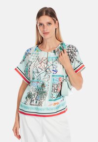Kurzarmbluse in Hellblau mit bunten Druckmotiven, schwarzen Umrandungen, roten und grünen Akzenten sowie einem dezenten Schlüsselloch-Ausschnitt.