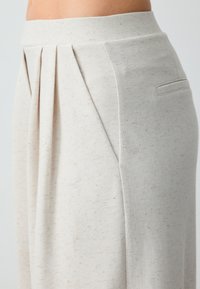 Pantalons beige clair et texturés avec des plis sur le devant, une taille lisse et une poche latérale discrète. Le tissu semble doux et décontracté.