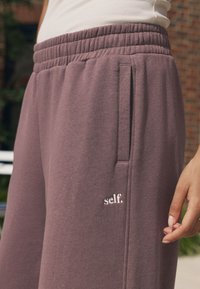 Pantaloni de trening mov cu talie elastică și buzunare laterale, având un mic logo alb "self." cusut în față. Textură moale.